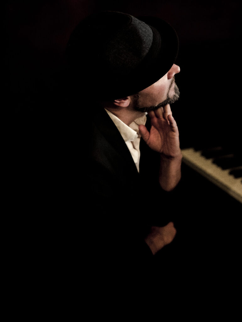 Pianist mit Hut sitzt nachdenklich am Klavier, Hand am Kinn, im dunklen Licht.