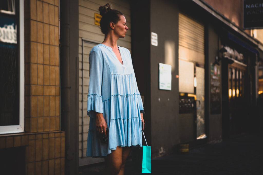 Frau in hellblauem Kleid steht mit Einkaufstasche in der Hand in einer Straße, das Gesicht zur Sonne gewandt.