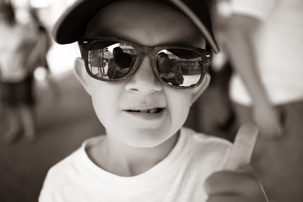 Junge mit Sonnenbrille zeigt Daumen hoch, in der Brille spiegelt sich der Fotograf mit Kamera.