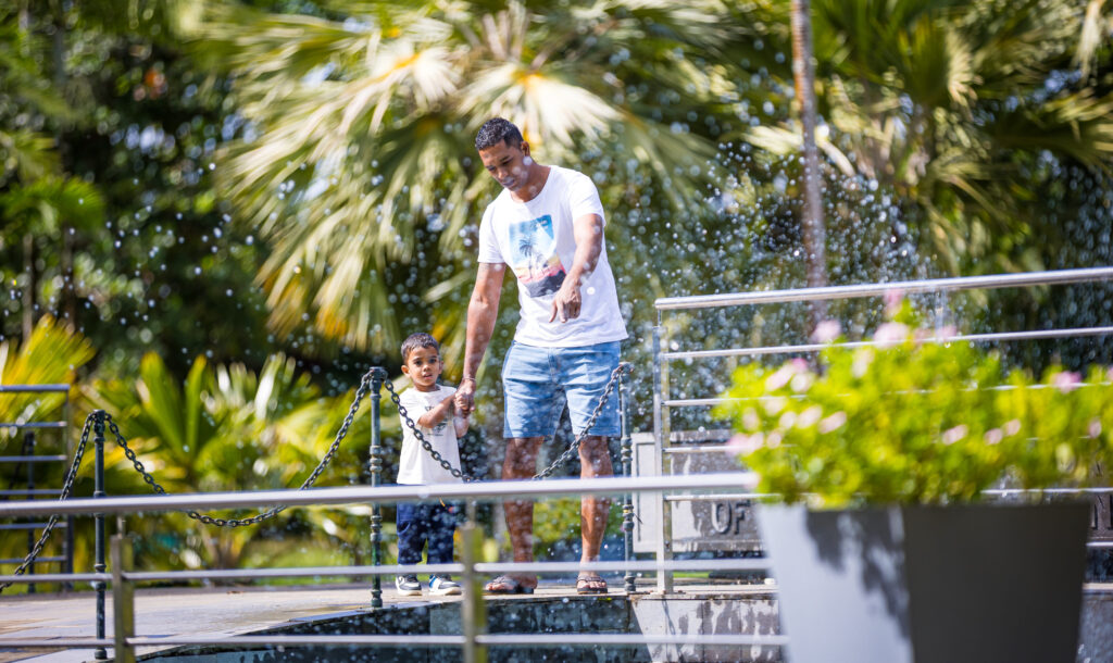 Ein Vater hält die Hand seines kleinen Sohnes und zeigt auf einen Brunnen, während Wasser in der Sonne sprüht.