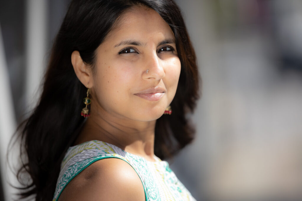 Frau mit dunklem Haar, feinen Ohrringen und Nasenpiercing blickt ruhig und selbstbewusst in die Kamera.