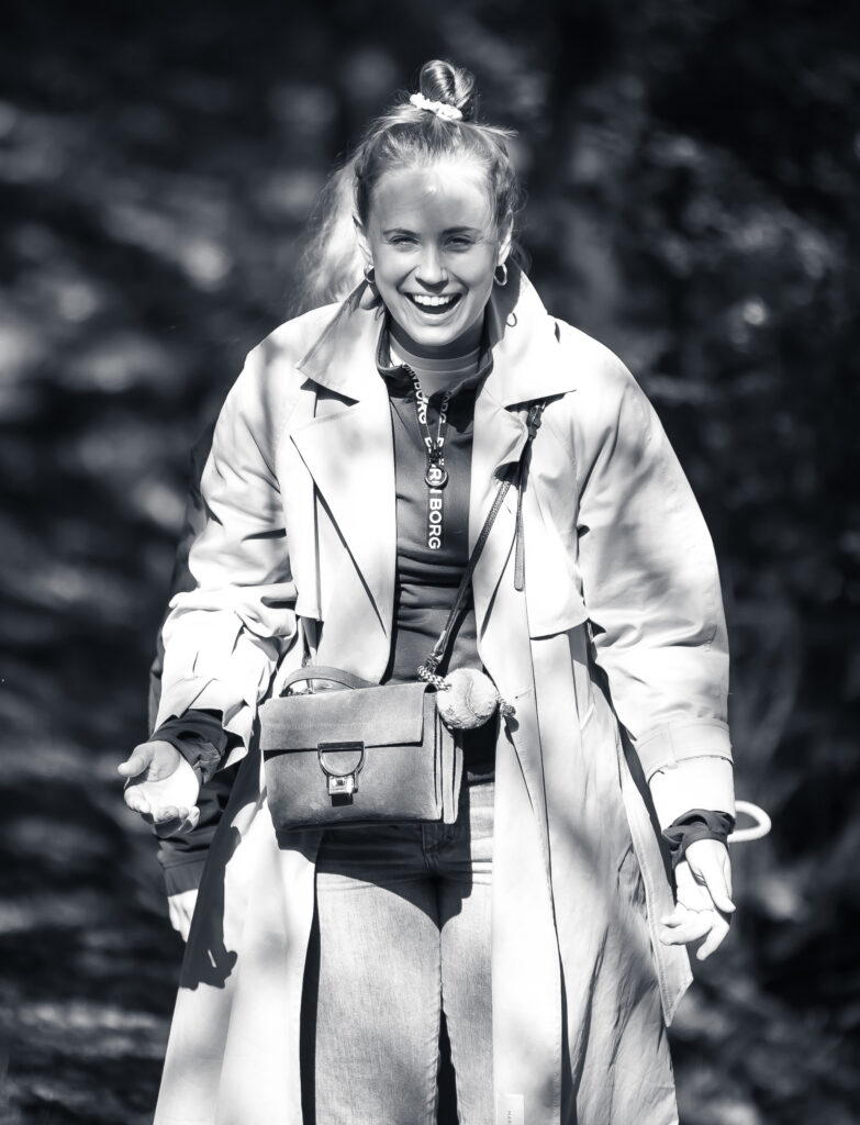 Junge Frau im Mantel lacht herzlich beim Spaziergang, Sonnenlicht fällt durch die Bäume.