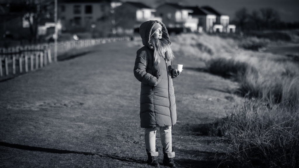 Frau im Wintermantel mit Tasse Kaffee in der Hand schaut in den Sonnenuntergang am Meer.