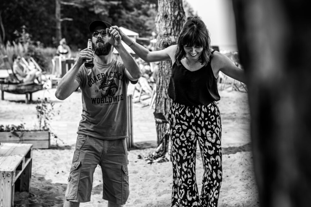 Ein Paar tanzt lachend am Strand, er mit Bierflasche in der Hand, sie in gemusterten Hosen voller Lebensfreude.