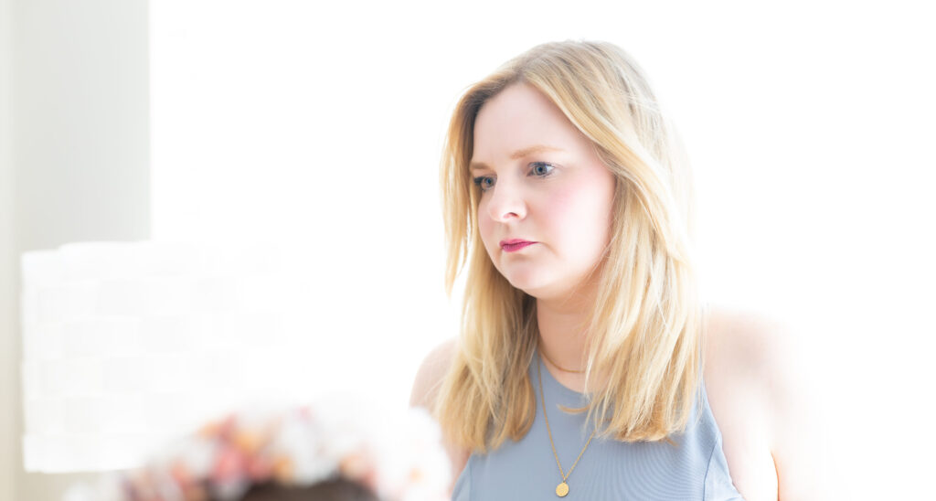 Junge Frau mit blondem Haar und grauem Top steht nachdenklich im hellen Licht, minimalistische Szene.