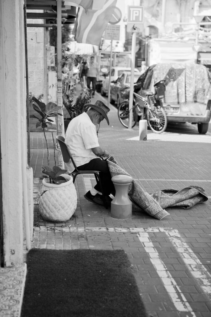 Älterer Mann mit Cowboyhut sitzt auf einem Stuhl und näht einen Teppich auf der Straße in Tel Aviv.