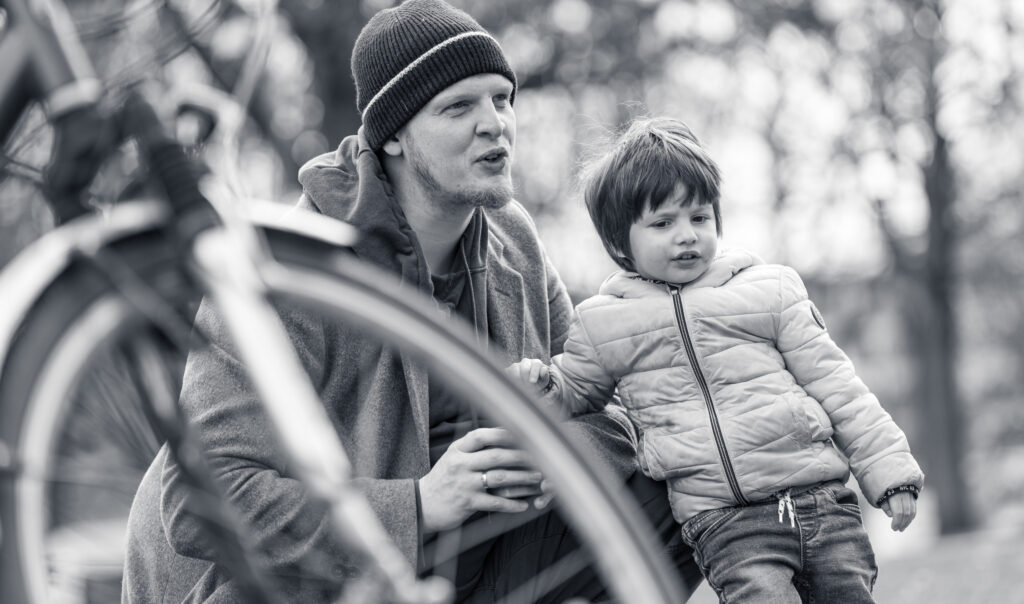 Vater mit Mütze sitzt neben seinem Kind im Park, ein Fahrrad ragt ins Bild.