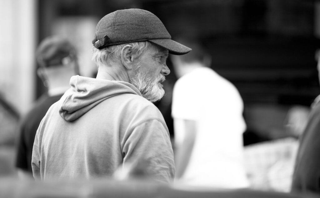 Älterer Mann mit Bart und Kappe in grauem Hoodie blickt nach unten, unscharfe Menschen im Hintergrund.