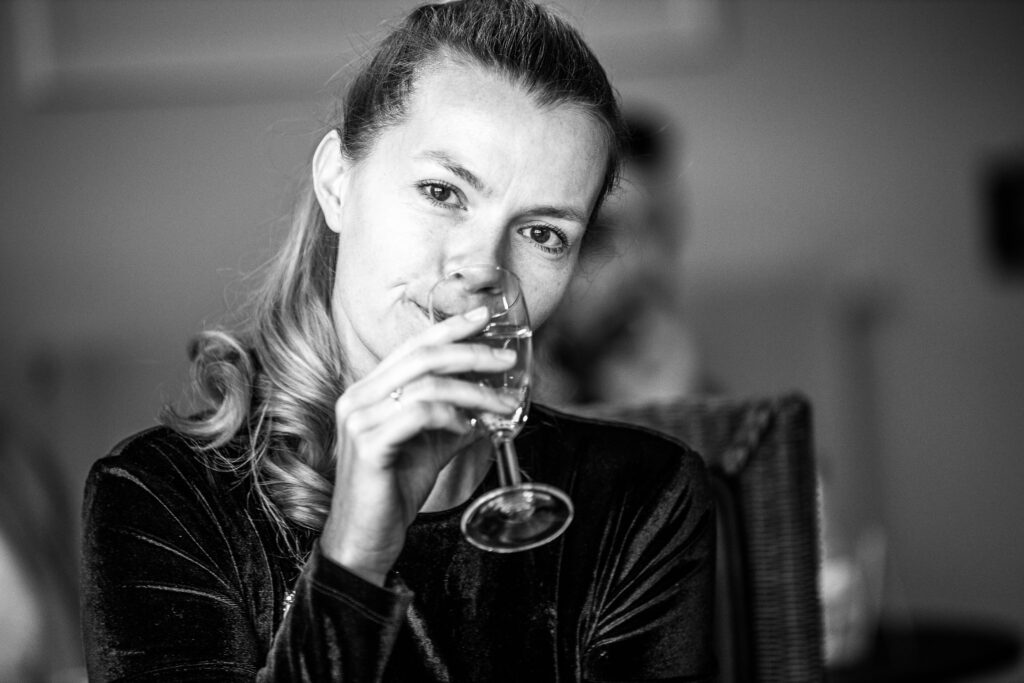 Frau mit zurückgebundenen Haaren hält ein Weinglas an den Mund und blickt direkt in die Kamera, unscharfer Hintergrund.