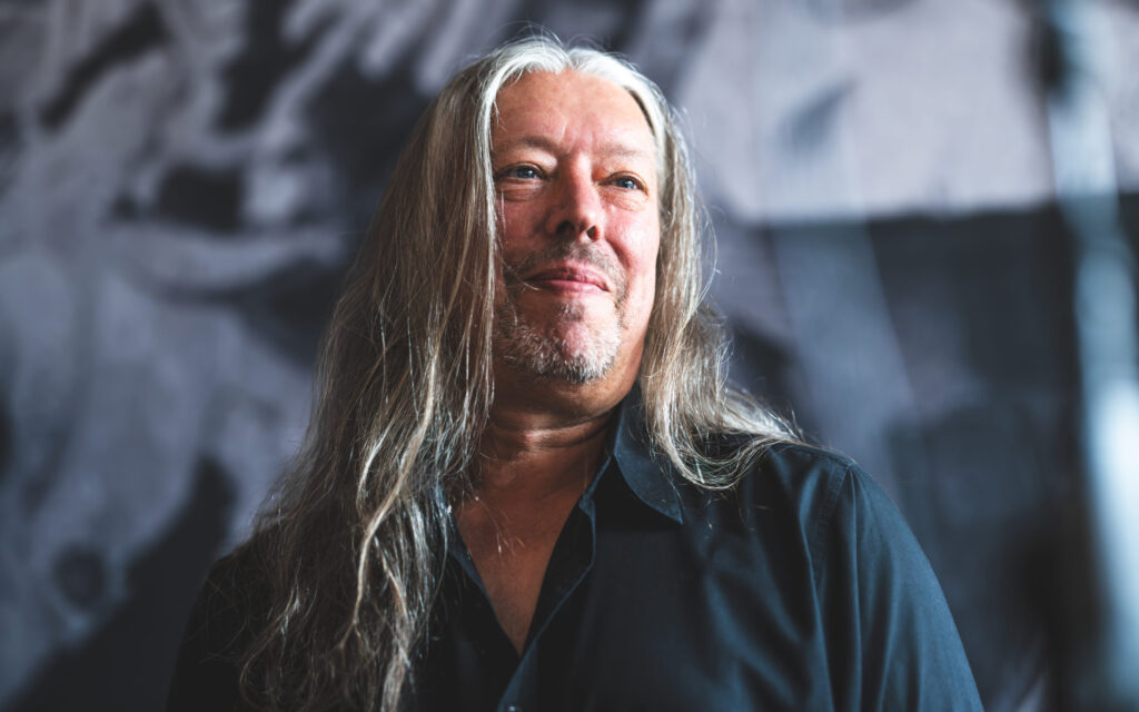 Porträt eines Mannes mit langen grauen Haaren und Bart, der lächelnd in die Ferne blickt, im Hintergrund verschwommene Strukturen.