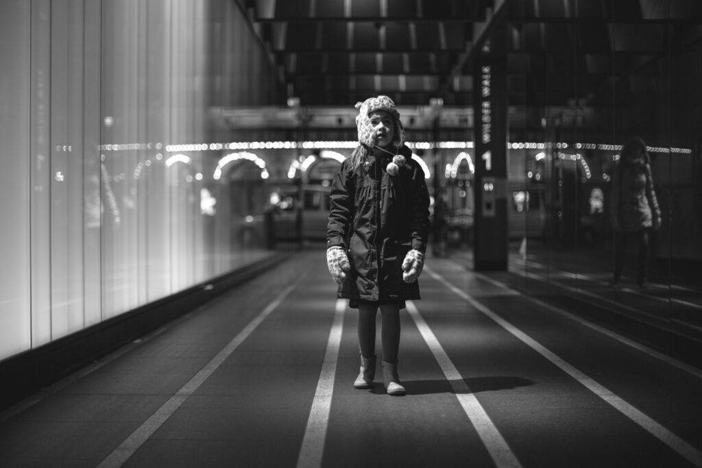 Ein Kind mit Winterjacke und Mütze steht nachts in einer hell erleuchteten Passage, im Hintergrund Lichter und Spiegelungen.