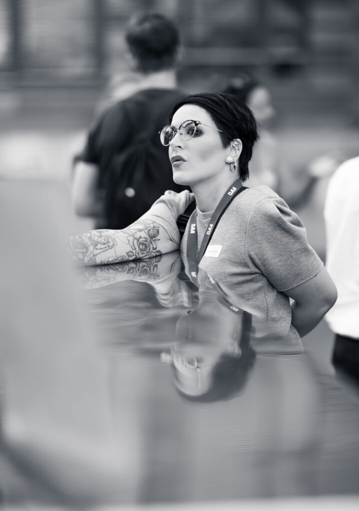 Frau mit Brille und Tattoos stützt sich nachdenklich an einer Fläche ab, unscharfe Menschen im Hintergrund.