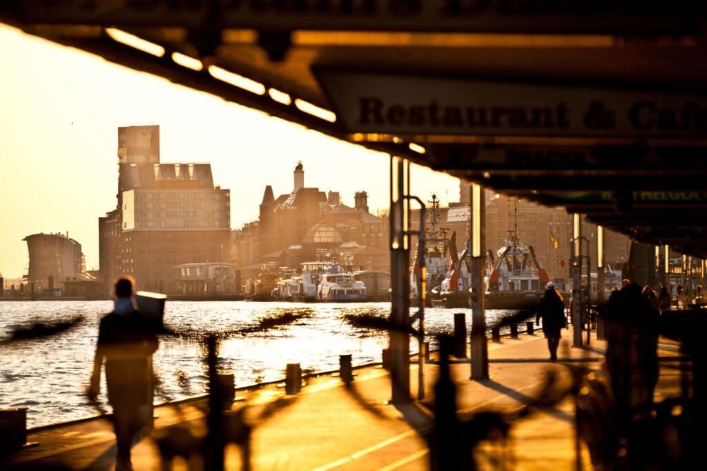 Abendstimmung am Hamburger Hafen, Spaziergänger am Wasser mit Blick auf Schiffe und den Fischmarkt im Sonnenlicht.