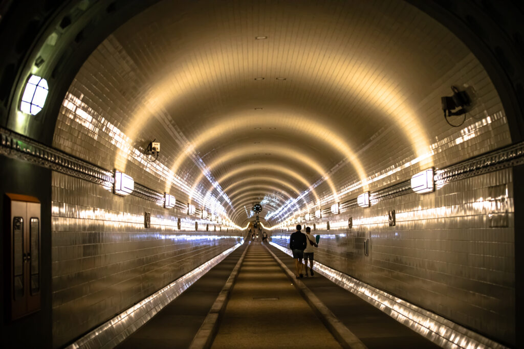 Beleuchteter Elbtunnel mit gekachelten Wänden und gewölbter Decke, Menschen spazieren durch den langen Gang.