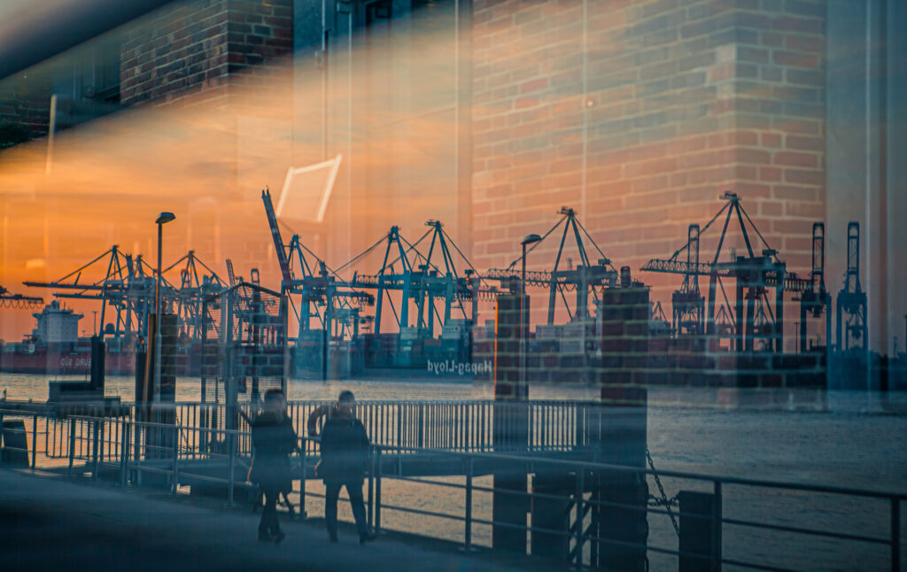 Spiegelung des Hamburger Hafens bei Sonnenuntergang mit Kränen und zwei Spaziergängern an der Elbe.