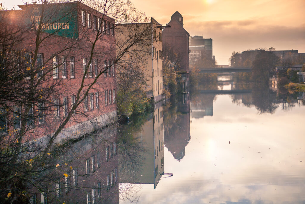 Rote und gelbe Backsteinbauten am ruhigen Kanal, Spiegelung im Wasser, Brücken im Hintergrund bei Sonnenaufgang.