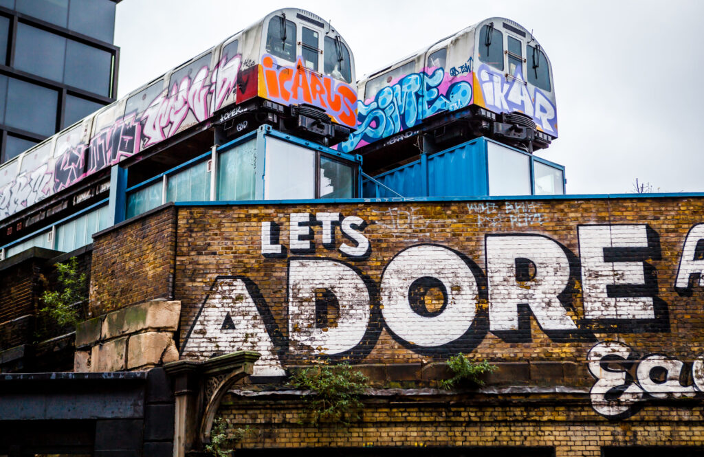 Graffiti-Straßenkunst mit alten U-Bahn-Wagen auf Containern und großem ‚LET’S ADORE‘-Schriftzug auf Backsteinmauer.