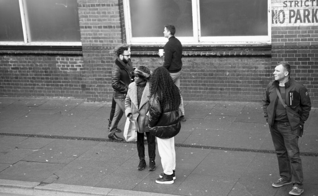 Straßenszene in Schwarz-Weiß mit fünf Passanten vor einer Backsteinwand, zwei Frauen im Gespräch im Vordergrund.