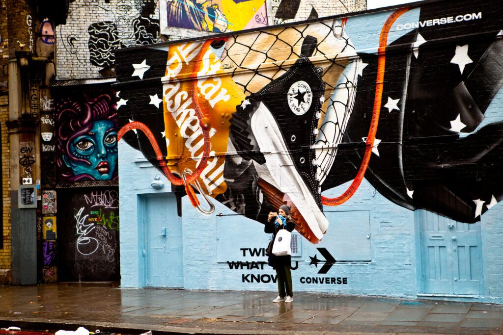 Bunte Streetart in Shoreditch mit großem Converse-Schuh, davor Frau mit Tasche fotografiert die bemalte Wand.