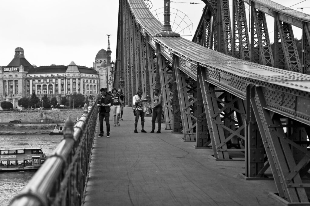 Fußgänger auf einer Stahlbrücke über die Donau in Budapest, im Hintergrund das Hotel Gellért mit markanter Kuppel.