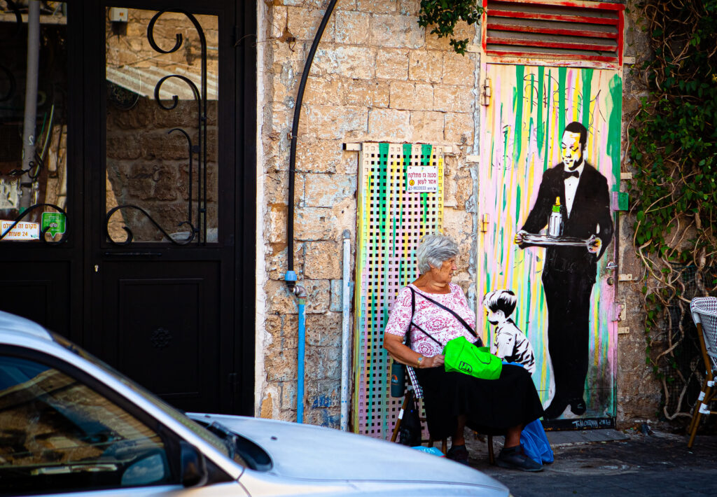 Ältere Frau sitzt in Tel Aviv vor Wand mit Streetart: Kellner mit Tablett und Kind, bunte Streifen im Hintergrund.