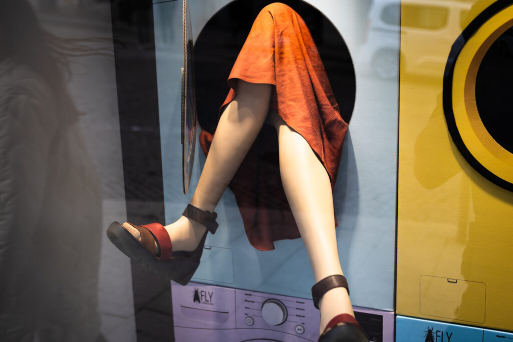 Schaufenster mit Schaufensterpuppe, deren Beine aus einer Waschmaschinentrommel ragen, neben bunter Werbung von Fly London.