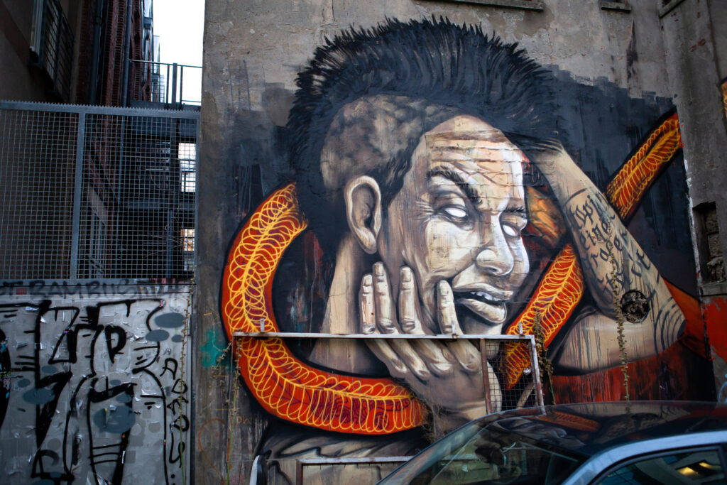 Großes Graffiti an Betonwand: Porträt einer Person mit weißen Augen, roter Schlange um den Hals, daneben Graffiti.