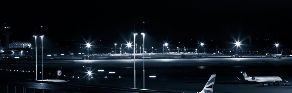 Nachtszene am Flughafen mit beleuchteten Start- und Landebahnen, parkenden Flugzeugen und hellen Scheinwerfern.
