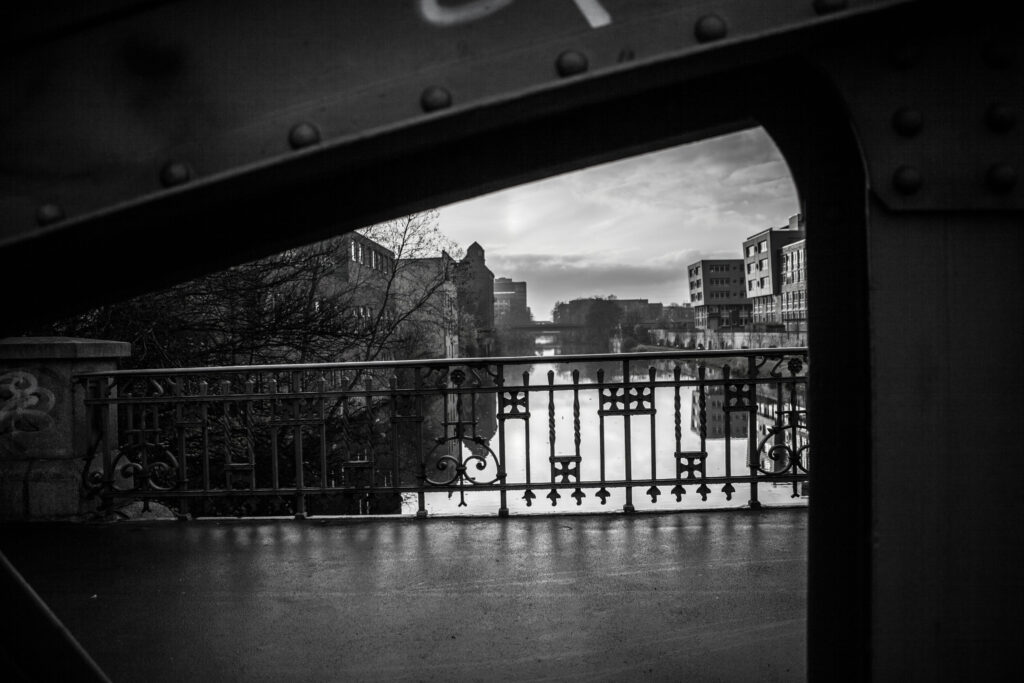 Stadtblick durch Stahlträger einer Brücke, Fluss mit Spiegelung, historische und moderne Gebäude im Hintergrund bei Dämmerung.