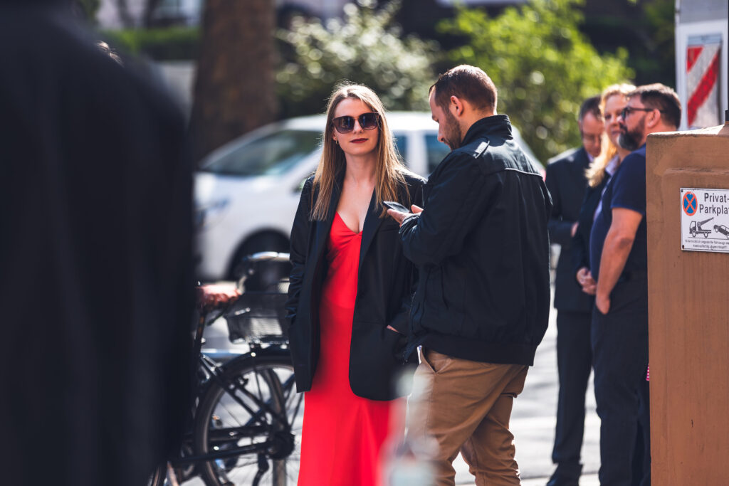 Frau im roten Kleid mit Sonnenbrille steht im Freien bei einer Veranstaltung, umgeben von anderen Gästen.