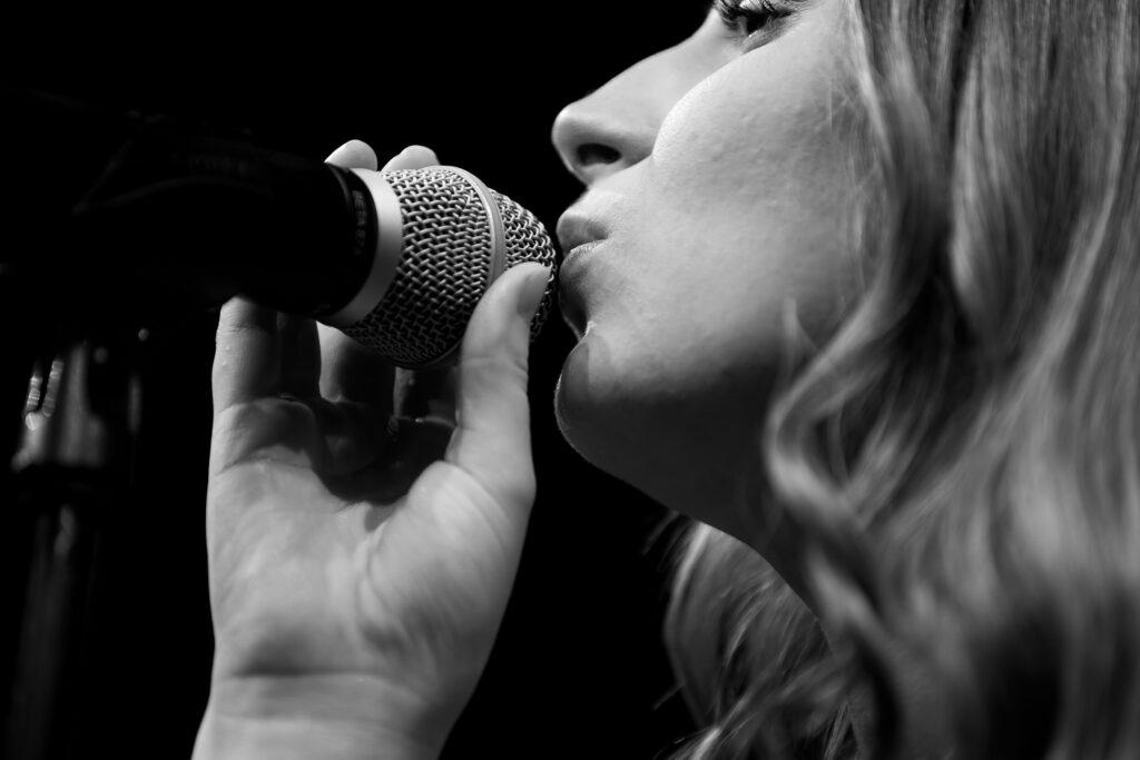 Schwarz-weiß Aufnahme einer Frau beim Singen, Mund dicht am Mikrofon, Nahaufnahme mit Fokus auf Lippen und Hand.