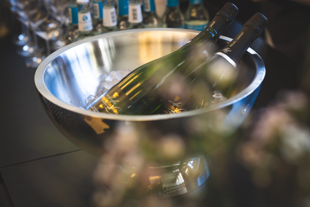 Zwei Flaschen Sekt in einer großen Metallschale mit Eiswürfeln, Gläser und Getränke im Hintergrund.