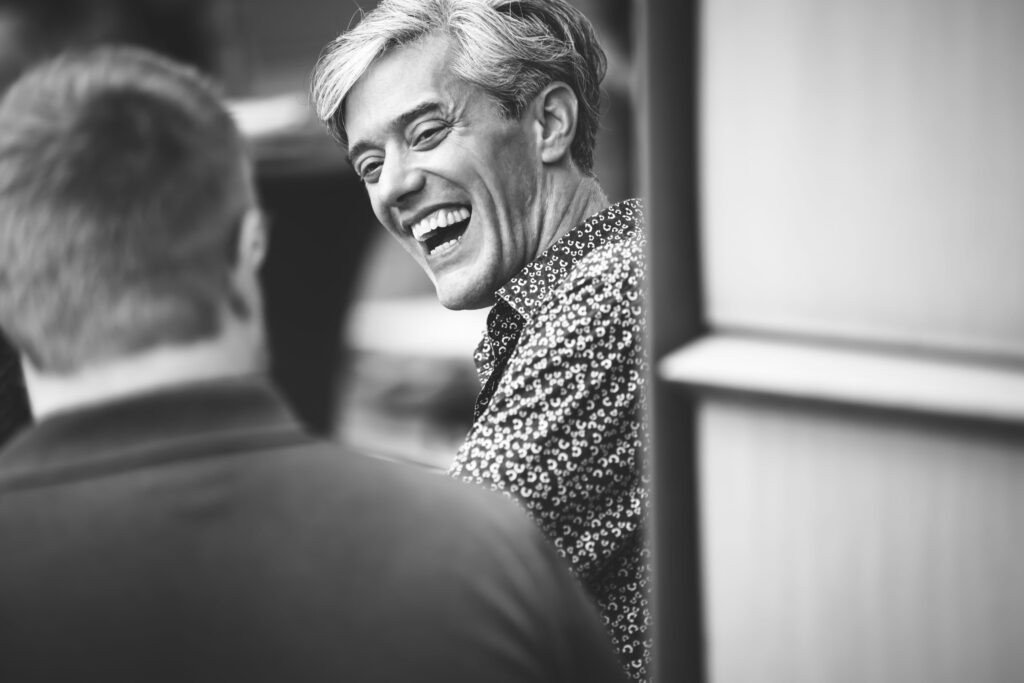 Mann mit gemustertem Hemd lacht herzlich im Gespräch, Schwarz-Weiß-Aufnahme voller Lebensfreude.