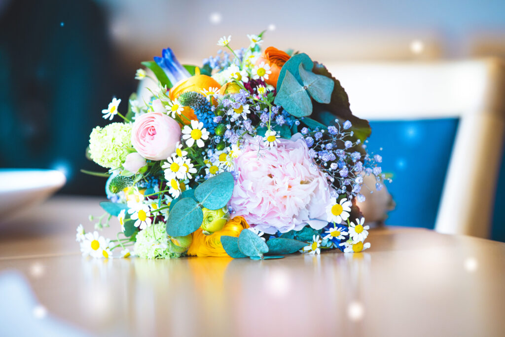 Bunter Blumenstrauß mit Rosen, Kamille und Pfingstrosen auf einem Tisch, im Hintergrund unscharfe Stühle.
