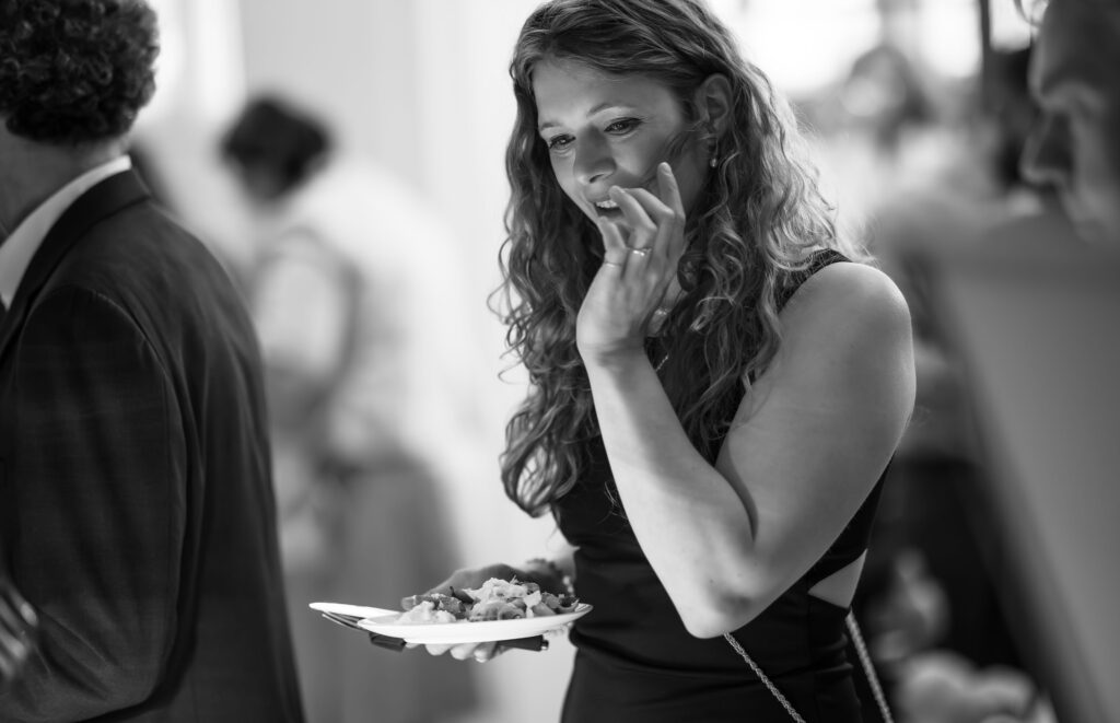 Frau mit lockigen Haaren hält einen Teller mit Essen, lächelt und hebt die Hand beim Gespräch.