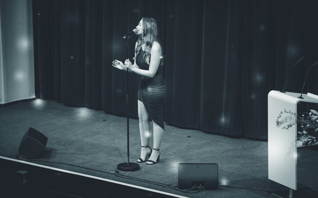 Frau im Kleid singt auf einer Bühne in ein Mikrofon, neben ihr ein Rednerpult, im Hintergrund ein geschlossener Vorhang.