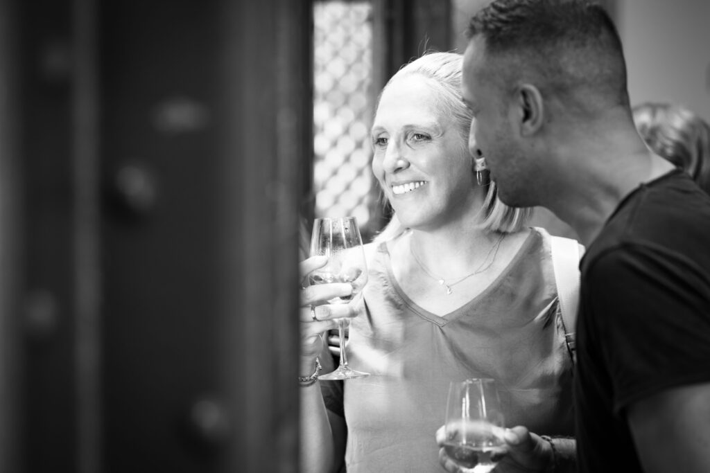 Frau und Mann stehen bei einer Veranstaltung beisammen, lächeln und trinken aus Weingläsern.
