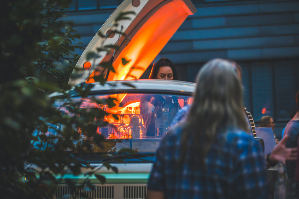 Frau schenkt Getränke an beleuchtetem Oldtimer-Bar bei einer Open-Air-Veranstaltung ein, Menschen im Vordergrund.