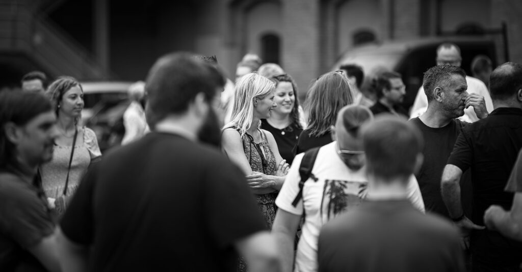 Schwarz-weiß-Aufnahme einer Menschengruppe im Freien, mehrere Personen im Gespräch, im Hintergrund Gebäude.