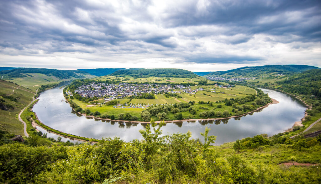 Moselschleife mit Weinbergen, grünem Tal und idyllischem Dorf unter bewölktem Himmel in Rheinland-Pfalz.