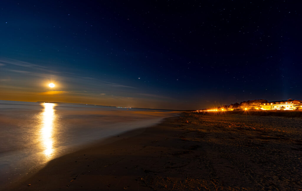 Nachts am Ostseestrand mit hellem Mondschein über dem Meer und leuchtenden Häusern am Ufer.