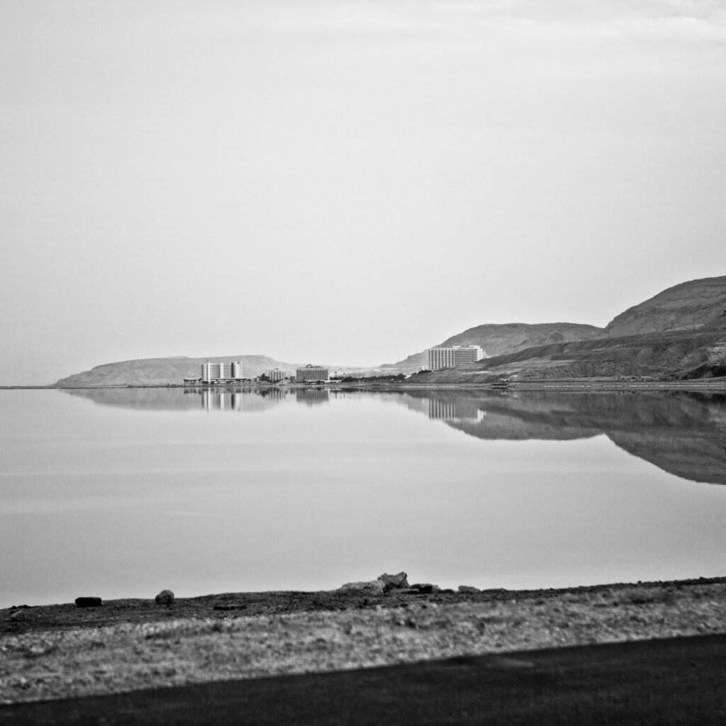 Schwarzweiß-Aufnahme vom Toten Meer mit spiegelglatter Wasserfläche, umliegenden Bergen und Hotels am Ufer.