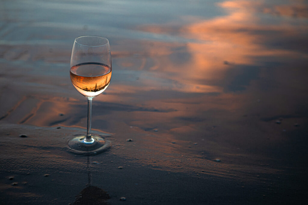Glas Roséwein im Abendlicht auf nassem Sandstrand, mit Spiegelung von orangefarbenen Wolken im Wasser.