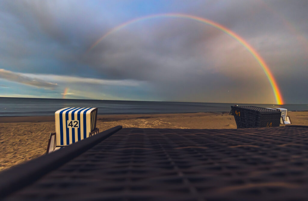 Strandkörbe am Ostseestrand mit Regenbogen über dem Meer und dramatischem Himmel nach einem Sommerregen.