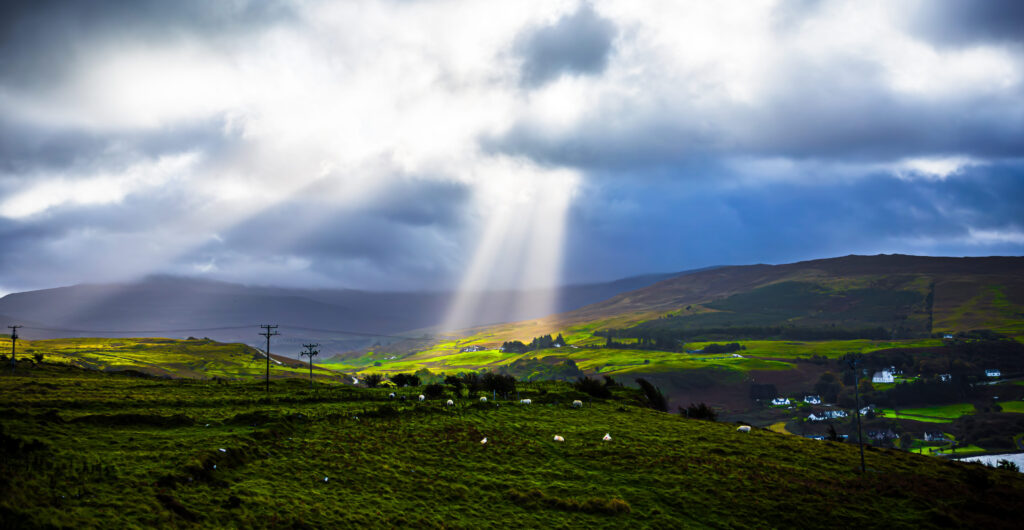 Sonnenstrahlen brechen durch Wolken über grünen Hügeln mit Schafen und Häusern in den schottischen Highlands.