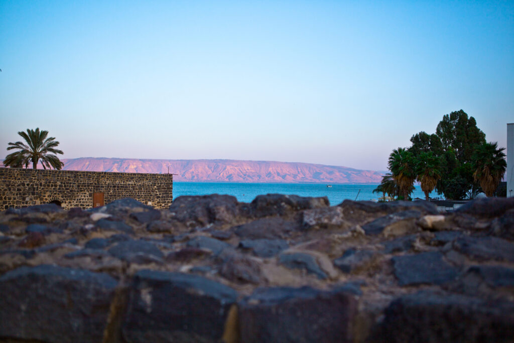Abendstimmung am See Genezareth mit Steinmauer, Palmen, blauem Wasser und rosa gefärbten Bergen im Hintergrund.