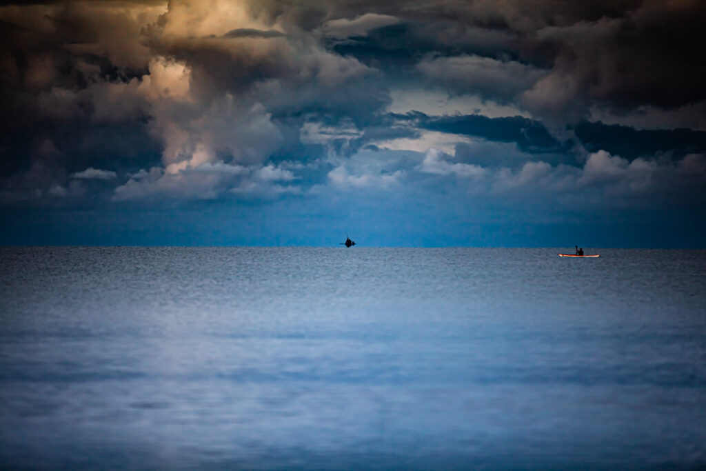Weite Meeresfläche unter dramatischem Himmel mit dunklen Wolken, kleines Segelboot und Kajak am Horizont.