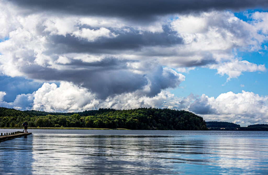 Dramatische Wolken über einem See mit grünem Waldufer, Steg und einsamer Person am Wasser.