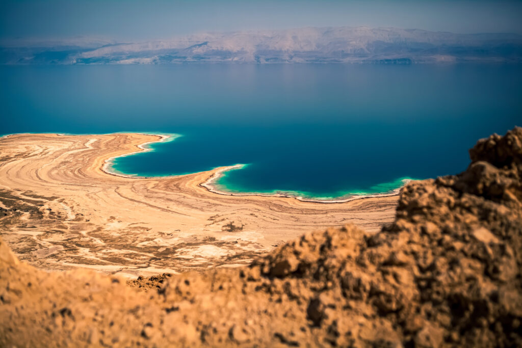 Küste des Toten Meeres mit türkisfarbenem Wasser, salzverkrustetem Ufer und Gebirgskette im Hintergrund.
