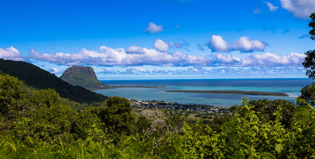 Blick auf Le Morne Brabant in Mauritius mit türkisfarbener Lagune, grünem Wald im Vordergrund und blauem Himmel mit Wolken.